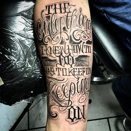 A forearm tattoo with the text "THE Only Thing I KNEW HOW TO DO WAS TO KEEP ON KEEPING ON" in ornate script. The arm rests on a black surface with a person seated in the background, symbolizing resilience often celebrated in tattoos St. Louis artists craft so well.