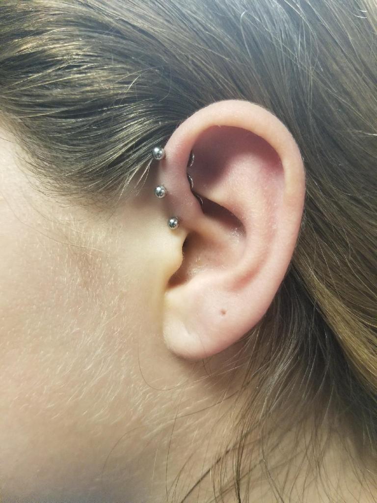 A close-up of a person's ear showing three silver ball piercings along the upper cartilage.