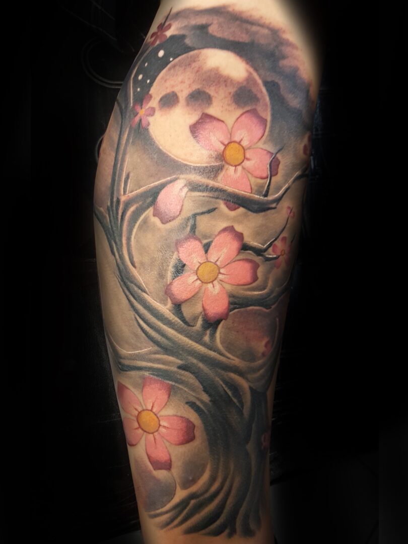 A tattoo on an arm depicts pink flowers on swirling branches with a moon and clouds in the background, capturing the enchanting artistry found in Tattoos St. Louis.